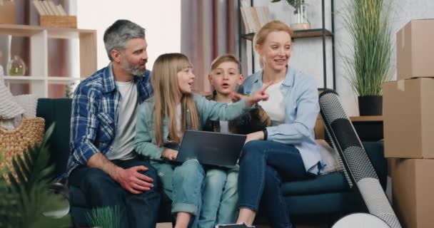 Atraente feliz família amigável positivo sentado no sofá em novo apartamento com um monte de caixas após a realocação e discutindo design usando o programa de computador — Vídeo de Stock