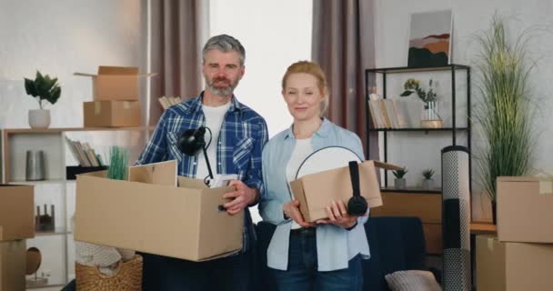 Conceito de realocação onde o casal adulto feliz sorridente bem-parecido está com caixas de papelão na frente da câmera em seu novo apartamento, vista frontal — Vídeo de Stock