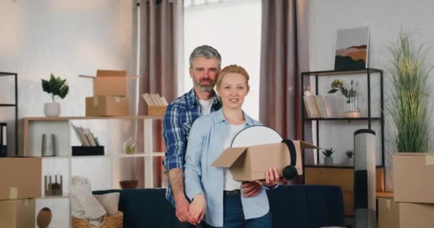 Adorable couple souriant heureux debout devant la caméra dans la chambre avec beaucoup de boîtes en carton et montrant les clés de leur nouvel appartement, vue de face — Video