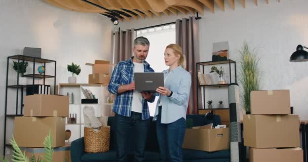 Probable couple marié confiant et de bonne humeur discutant d'idées pour meubler leur nouvelle maison, ils utilisent une application spéciale sur ordinateur portable lorsqu'ils se tiennent près des boîtes en carton environnantes dans la chambre — Video