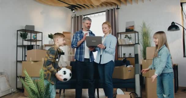 Roztomilý šikovný spokojený dospělý pár diskutovat nápady na design místnosti pomocí počítačového programu, zatímco jejich veselý hravý 10-ti letý syn a dcera baví s míčem — Stock video