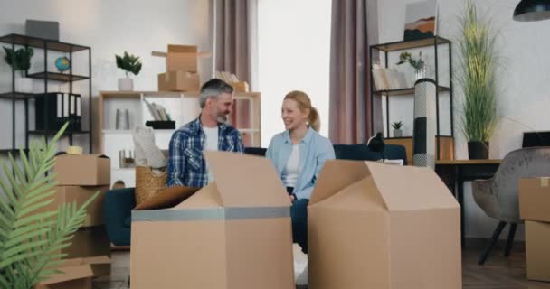 Boa aparência feliz casal adulto despreocupado positivo desfrutando de conversa conjunta no novo apartamento quando seus brincalhão alegre dois filhos pulando para fora de caixas de papelão e acenando as mãos — Vídeo de Stock