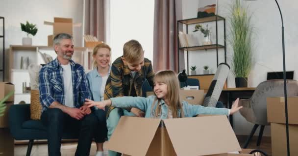 Attraente sorridente genitori felici amorevoli guardando come gioire due bambini, figlia seduta in scatola di cartone e fare movimenti volanti mentre il figlio la spinge in scatola — Video Stock