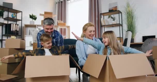Счастье в новом доме, где красивые веселые беззаботные родители играют со своим сыном и дочерью, которые сидят в коробках, пока мама и папа толкают их — стоковое видео