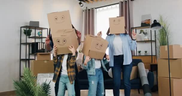 Семейное времяпрепровождение в новой квартире, где неузнаваемо веселая семья с двумя детьми, надевая коробки на голову, танцует в еще не полностью меблированной комнате — стоковое видео