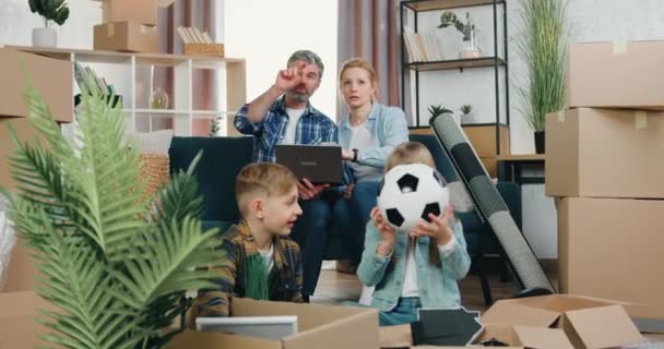 Snygga positiva moderna par sitter på soffan och diskuterar design av sin nya lägenhet med hjälp av datorprogram medan barn packa upp kartonger, omlokalisering koncept — Stockvideo