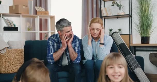 Устал расстроенный бородатый мужчина и блондинка сидят на диване и смотрят, как радуются их два ребенка-сына и дочь среди коробок, потому что они переезжают в новую квартиру — стоковое видео