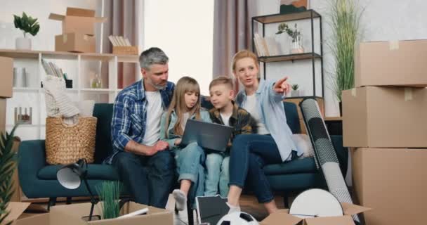 Concepto de reubicación donde una atractiva familia feliz y moderna se sienta en el sofá entre cajas de cartón y discute el diseño de la habitación usando el programa de computadora — Vídeos de Stock
