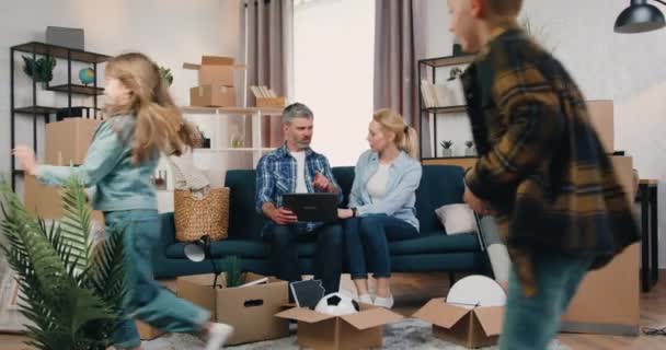 La gente y el concepto de bienes raíces donde atractiva pareja segura positiva sentado en el sofá en el nuevo apartamento y discutir el diseño de la habitación, mientras que sus dos hijos jugando alrededor de cajas de cartón — Vídeo de stock