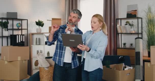 Attrayant heureux couple moderne positif debout au milieu de la pièce dans le nouvel appartement et discuter de la conception à l'aide d'un programme spécial sur ordinateur — Video