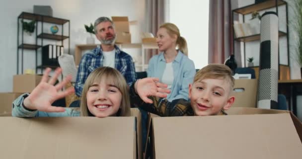 Heerlijk tevreden vrolijke jongen en meisje zitten in kartonnen dozen en zwaaien handen in de camera terwijl hun positieve ontspannen ouders zitten op de bank en praten tussen elkaar — Stockvideo