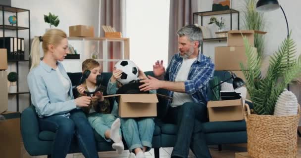 Atractiva familia moderna positiva y amigable sentada en el sofá en un apartamento nuevo después de la reubicación, los niños ayudan a los padres a desempacar cajas de cartón con diferentes cosas pequeñas y agradables. — Vídeo de stock