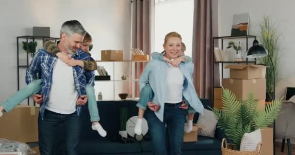Goed uitziende glimlachende zorgeloze ouders hebben plezier samen met hun zoon en dochter van 10 jaar oud, dragen ze achter de rug en draaien met hen in een nieuw huis — Stockvideo
