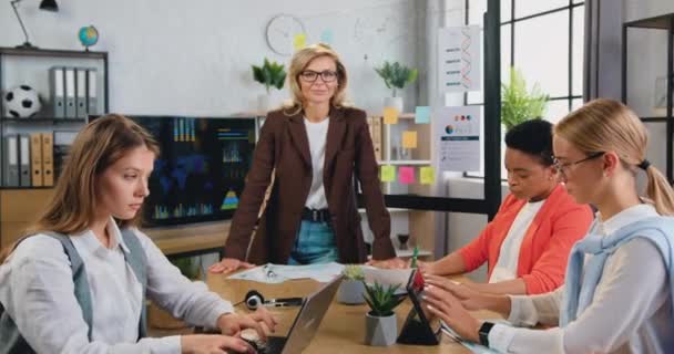 Front view of beautiful adult leader woman in glasses, standing in modern office looking at camera, with team of mixed race women working successfully at briefing table in office — Stock Video