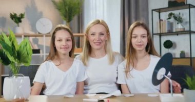 Çekici, neşeli, mutlu, dost canlısı sarışın anne ve iki genç kızı kameranın önünde oturuyor. Güzel dekore edilmiş oturma odasında samimi bir şekilde gülümsüyorlar.