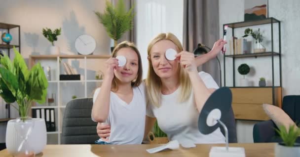 Aantrekkelijke glimlachende speelse blonde vrouw met tienermeisje zetten op hun ogen katoen pads en dan het nemen van hen terwijl kijken in de camera in gezellige interieur — Stockvideo