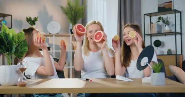 Portrét dobře vypadající vtipné, šťastně se usmívající blond matky a jejích dospívajících dcer, které si doma užívají společný volný čas, sundávají si z očí půlku citronu, pomeranče a grapefruitu a smějí se — Stock video