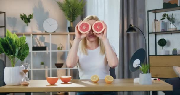 Krásná usměvavá veselá hravá blondýnka pózující na kameru s půlkou grapefruitu na jejích očích na současném domácím interiéru pozadí — Stock video