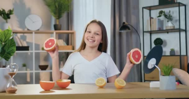 Roztomilý šťastný usměvavý teen maják dívka sedí před kamerou doma a hraje s polovinou grapefruit, přední pohled, zpomalený film — Stock video