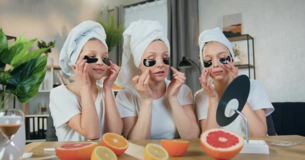 Schattige glimlachende blije zorgzame moeder en haar twee 12-14-jarige dochters alle in badstof handdoeken op hun hoofd zitten in de voorkant van kleine spiegel thuis, terwijl onder ogen verfrissende pleisters — Stockvideo