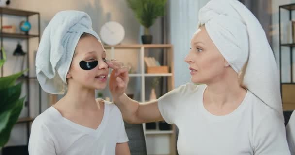 Härligt positiv omtänksam kärleksfull mor och hennes tonåring dotter i frotté handdukar runt huvudet tillämpa uppfriskande kollagen fläckar under ögonen, hudvård koncept — Stockvideo
