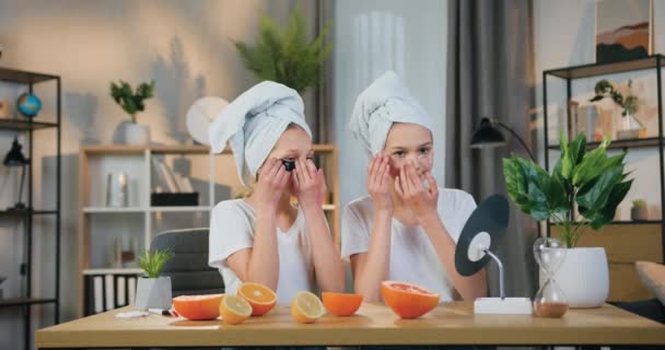 Cute friendly pozytywne dwie nastolatki z ręcznikami frotte na głowach siedzi przed lustrem w przytulnym salonie w domu i stosowania kolagenu opaski na oczy — Wideo stockowe