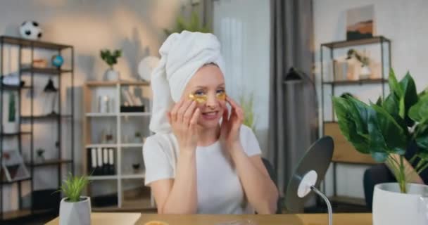 Camera schieten op mooie glimlachende gelukkige vrouw in witte handdoek op haar hoofd die het aanbrengen van gouden collageen oogvlekken en kijken in de camera met vriendelijke gezichtsuitdrukking — Stockvideo