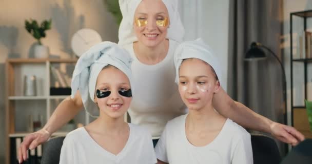 Hermosa madre feliz sonriente amigable y dos hijas adolescentes con toallas de felpa en la cabeza y con refrescantes parches de colágeno en la cámara en la acogedora sala de estar — Vídeos de Stock