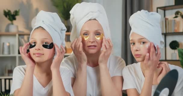 Splendide famille amicale souriante et heureuse comme mère et adolescentes dans des serviettes éponge autour de leur tête posant sur la caméra avec des patchs hydrogel yeux naturels, vue de face — Video