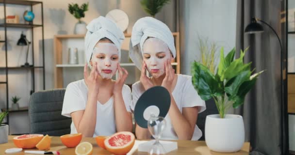 Dobrze wyglądający szczęśliwy pozytywne dwie nastolatki w ręczniki frotte wokół ich głowy stosując kosmetyczne maski twarzy siedzi przed lustrem w przytulnym salonie — Wideo stockowe