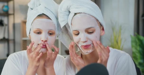 Primer plano retrato de atractiva sonrisa satisfecha amigable dos hermanas adolescentes con toallas alrededor de las cabezas que hacen procedimientos cosméticos faciales en casa — Vídeo de stock