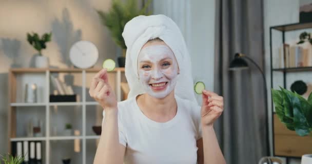 Ansiktsbehandling hudvård koncept där vackra leende glad kvinna med handduk runt huvudet tillämpa ansiktsmask och sätta gurka skivor på ögonen tittar in i kameran — Stockvideo