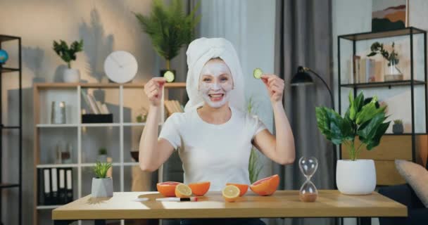 Imádnivaló boldog vidám modern felnőtt nő törölközővel a feje körül pózol a kamera közben, hogy arc eljárások tejszín és uborka szeletek, elölnézet — Stock videók