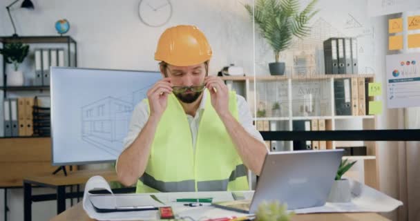 Architecte barbu confiant attrayant dans le casque et gilet assis à la table dans le bureau de conception et de travailler avec le plan de papier, concept de flux de travail — Video