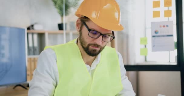 Close-up van sympathieke geconcentreerde doelgerichte bebaarde architect in vest en helm in bril die werkt met blauwdruk in gezellig design kantoor — Stockvideo