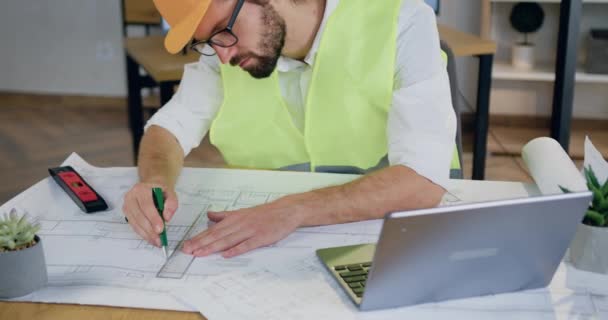 Sympathisch konzentrierter, bärtiger Konstrukteur in Helm und Weste zeichnet mit Stift und Lineal in gemütlichem Designbüro Linien auf Papierplan — Stockvideo