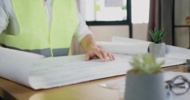 İyi görünümlü, deneyimli, kasklı, yelekli, kasklı ve sakallı bir mimar atölyedeki masada oturuyor ve taslağı inceliyor.