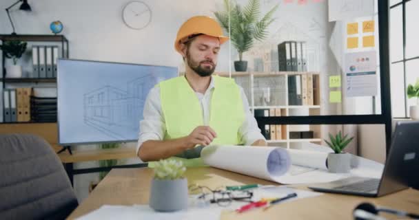 Atrakcyjny, pewny siebie, doświadczony, brodaty inżynier w kasku i kamizelce, siedzący przy stole w biurze projektowym i realizujący papierowy plan studiów, koncepcja przemysłu budowlanego — Wideo stockowe