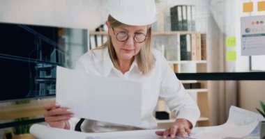 İyi görünümlü, konsantre olmuş, maksatlı yetişkin kadın mühendis miğfer kontrolü raporları ve modern tasarım ofisindeki geleceğin planlarını hazırlıyor.