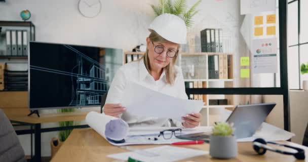 Bela confiante de alta qualificação de 45 anos loira principal arquiteto feminino em hardhat comparando dados com informações sobre computador, relatórios e esboços desenhados no escritório de design — Vídeo de Stock