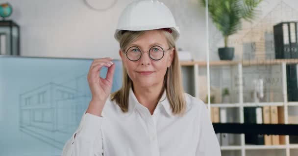 Atraente sorrindo bem sucedido profissional adulto engenheiro principal feminino no capacete tirando os óculos enquanto olha para a câmera no escritório de design moderno, de perto — Vídeo de Stock