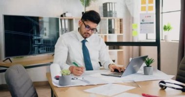 Ofis işi konsepti. Çekici, sorumluluk sahibi, sakallı ve gözlüklü bir işçinin dizüstü bilgisayardaki bilgilerle veri karşılaştırması üzerine notlar yazması.