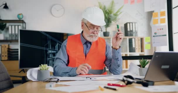 Likable Przemyślany profesjonalny szanowany brodaty inżynier w twardej czapce i kamizelce pracy z planu i akcesoriów rysunkowych w nowoczesnym biurze inżynieryjnym, widok z przodu — Wideo stockowe