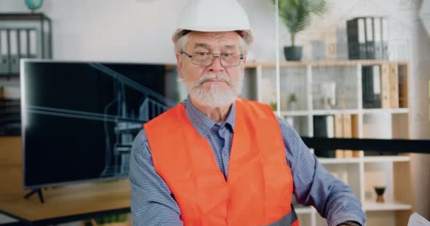 Närbild porträtt av snygg positiv säker professionell mogen skäggig byggarbetare i hjälm och väst som tar av sig glasögonen medan du tittar in i kameran i designkontoret — Stockvideo