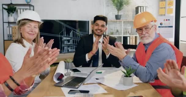 Attrayant souriant satisfait équipe multiethnique expérimentée de spécialistes de la construction dans les casques et gilets applaudissements mains après la fin réussie du projet commun dans la salle de réunion — Video