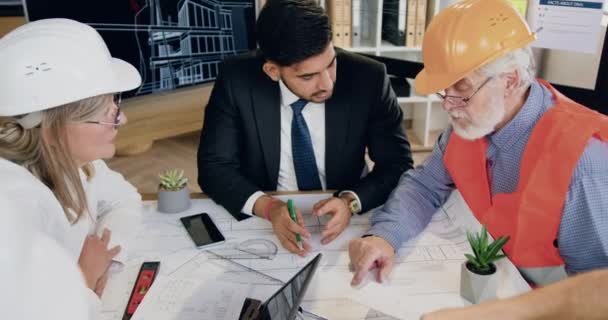 Attrayant confiant hautement qualifié diversifié équipe masculine et féminine de travailleurs de la construction en uniforme spécial remue-méninges sur le plan de construction dans le bureau d'études moderne — Video