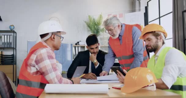 Mühendis-inşaatçılardan oluşan, başarılı sakallı ana mühendis ile birlikte toplantı odasındaki inşaat projesi üzerinde beyin fırtınası yapan başarılı, yetenekli bir ekip. — Stok video
