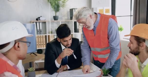 Çekici, sakallı, olgun bir inşaatçının ekibin diğer üyeleriyle yaptığı ortak toplantıda deneyimli bir mühendise plan detaylarını açıkladığı tasarım ofisinde çalışmak. — Stok video