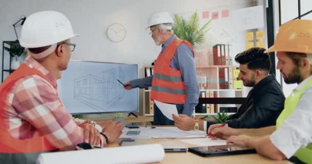 Knap positief vertrouwen ervaren senior bebaarde ingenieur in helm en vest presenteren bouwproject op tv scherm voor succesvolle professionele mannelijke en vrouwelijke collega 's — Stockvideo