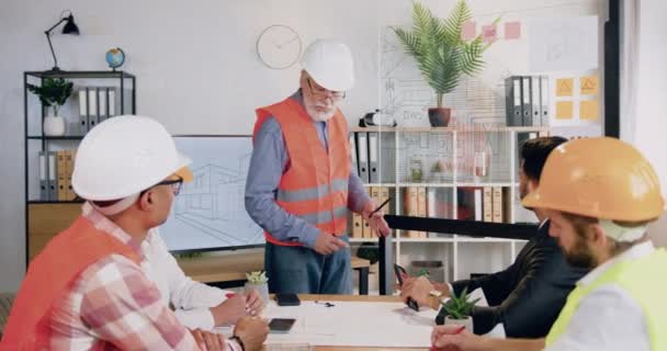 Un bel ingénieur barbu expérimenté et confiant dans le casque et le gilet expliquant le projet de construction dépiqué sur l'affichage numérique et le mur de verre pour ses divers collègues dans la salle de réunion — Video